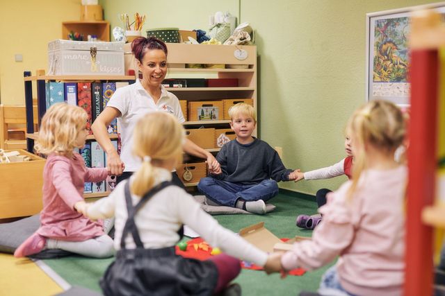 Kinder und Erzieherin sitzen im Kreis und fassen sich an die Hände.