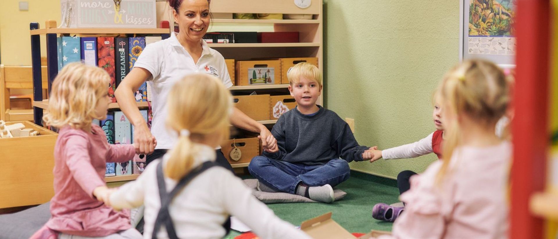 Kinder und Erzieherin sitzen im Kreis und fassen sich an die Hände.