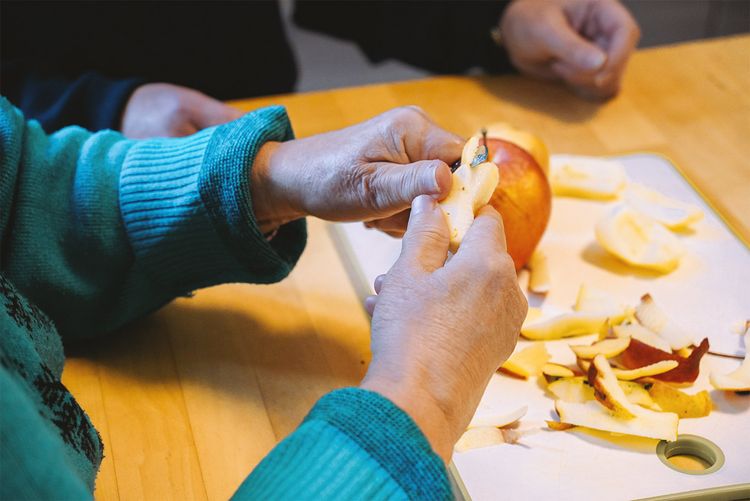 Die Hände einer Nutzerin des Dienstes schälen einen Apfel mit Unterstützung der Fachkraft.