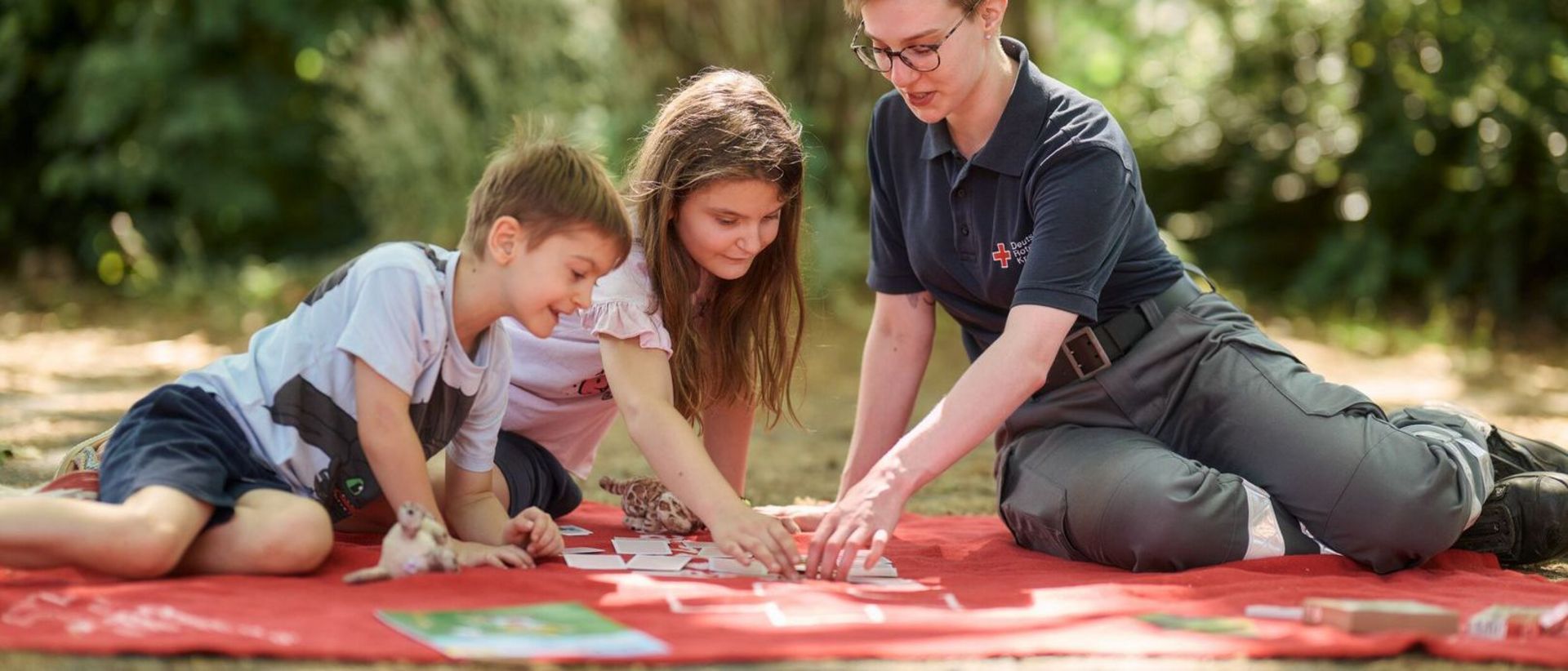 Kinder und DRK-Mitarbeiterin spielen im Garten ein Kartenspiel.