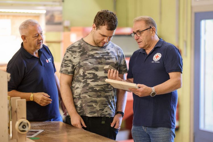 Zwei Mitarbeiter erklären einem Nutzer eine Arbeitsaufgabe am Werkstück Holz.
