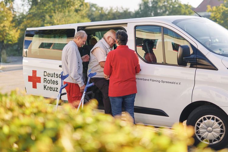 DRK Mitarbeiter unterstützen einen Herren beim Aussteigen aus dem Fahrdienst-Fahrzeug.