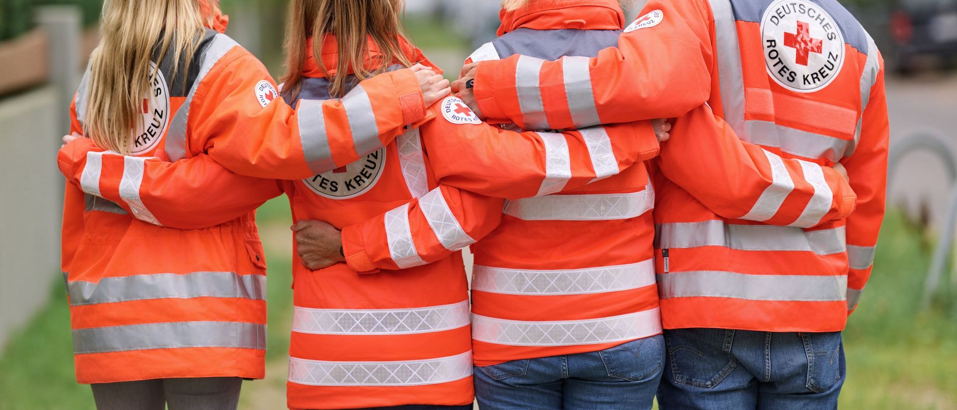 Rückansicht von Menschen in DRK-Einsatzjacke, welche durch die Geste des Umarmens Zusammenhalt vermitteln.