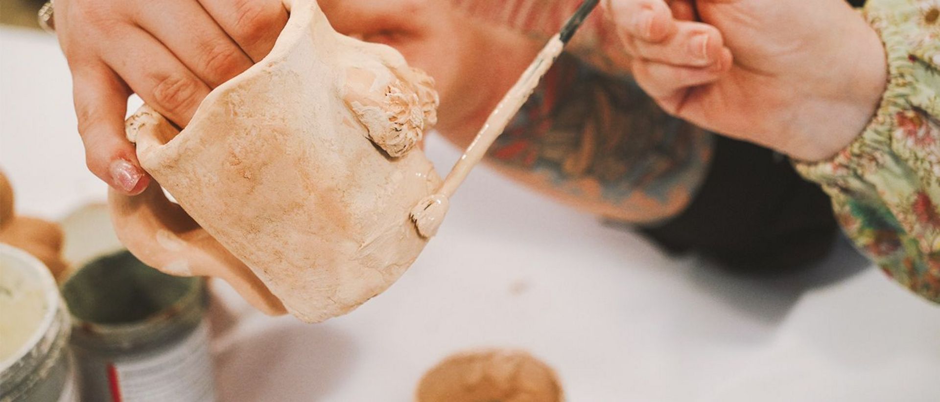 Man sieht Hände, welche eine Tasse mit einem Pinsel bemalen und welche dabei assistieren.