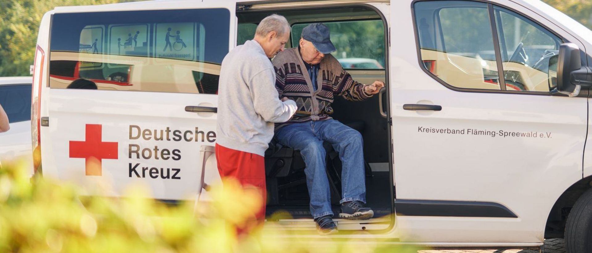 Auf diesem Foto sehen Sie einen großen Pkw, aus welchem ein Mitarbeiter des DRK Fahrdienstes einen Insassen beim Aussteigen hilft.