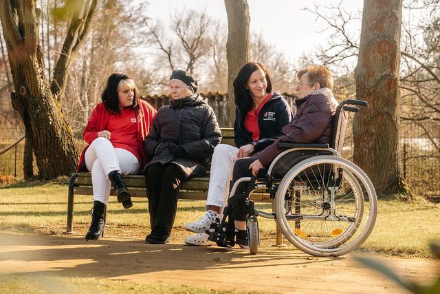 Mitarbeitende und Bewohnende Senioren einer Pflegeeinrichtung sitzen im Park auf einer Bank.