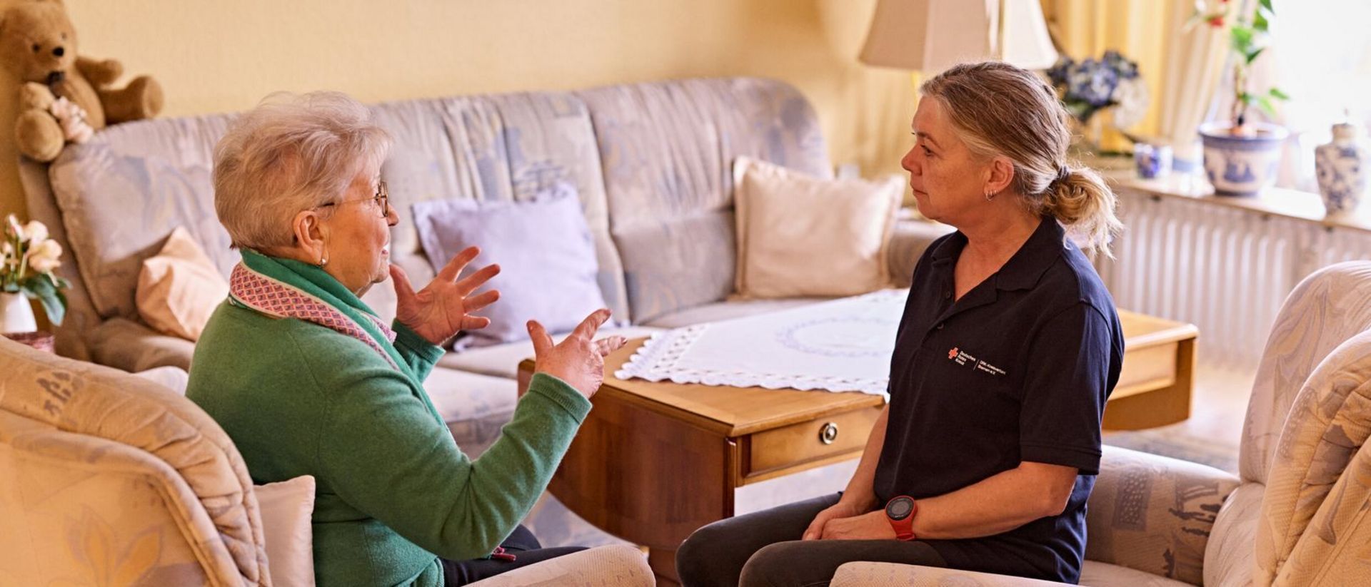 DRK-Mitarbeiterin sitzt gemeinsam mit Seniorin auf der Couch im Wohnzimmer.