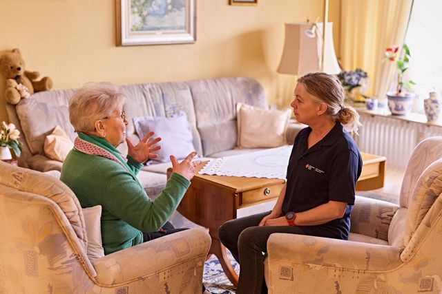 DRK Mitarbeiterin und Seniorin sitzen im Wohnzimmer bei nettem Gespräch.
