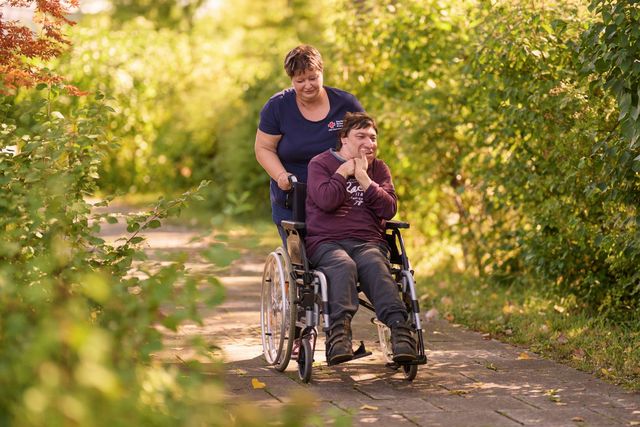 Mitarbeiterin schiebt Nutzerin im Rollstuhl durch einen Park.