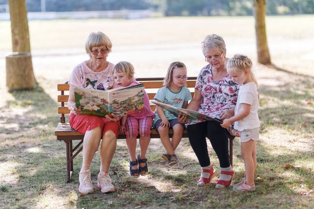 Zwei ältere Damen sitzen auf einer Parkbank mit Kindern und lesen gemeinsam Bücher.