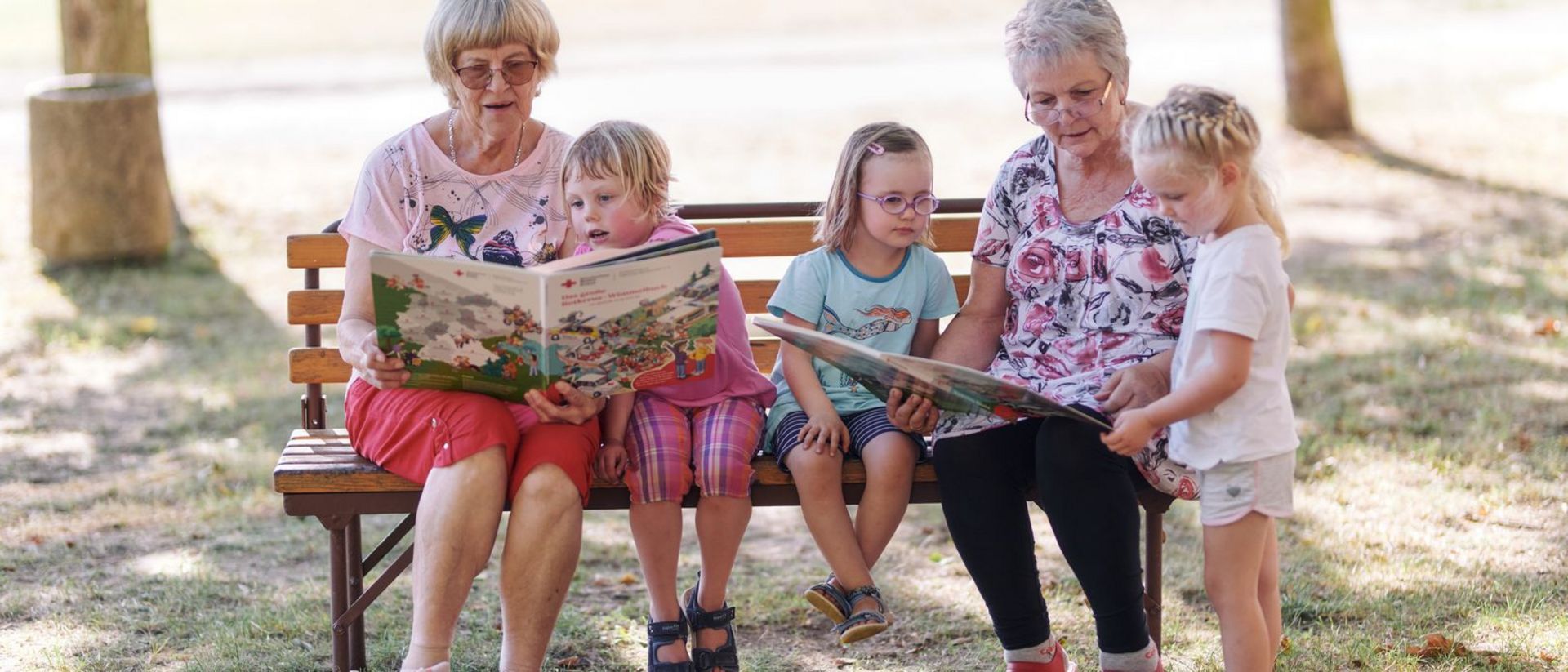 Zwei ältere Damen sitzen auf einer Parkbank mit Kindern und lesen gemeinsam Bücher.