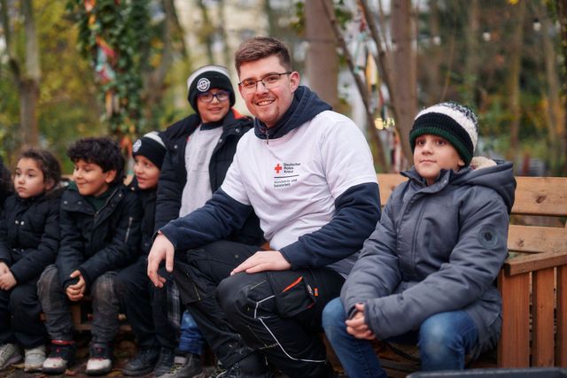 DRK Mitarbeiter sitzt mit Schulkindern auf einer Bank.