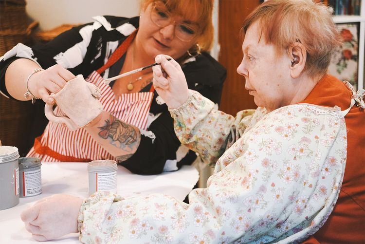 Eine Nutzerin bemalt mit Unterstützung einer Mitarbeiterin ein getöpfertes Werk.