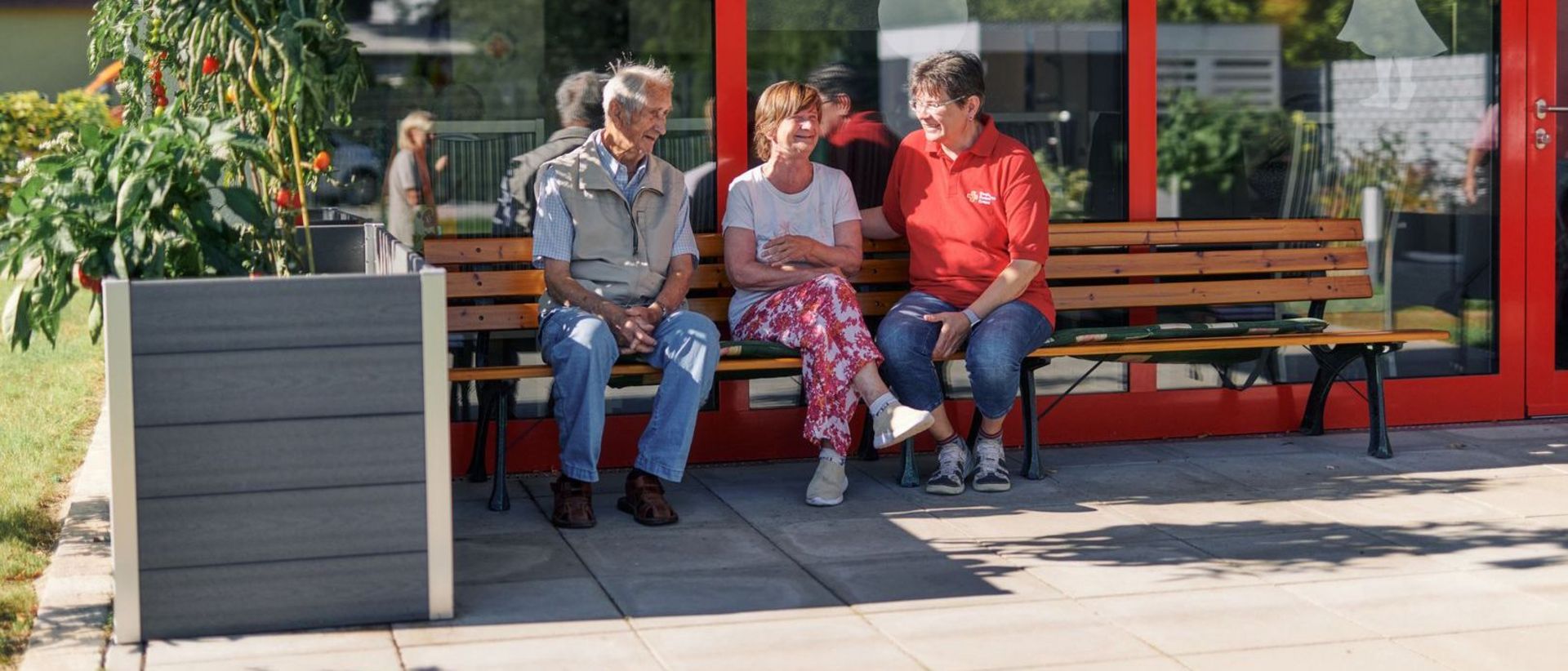 Mitarbeiterin des DRK sitzt mit Gästen der Tagespflege auf einer Bank im Garten. 