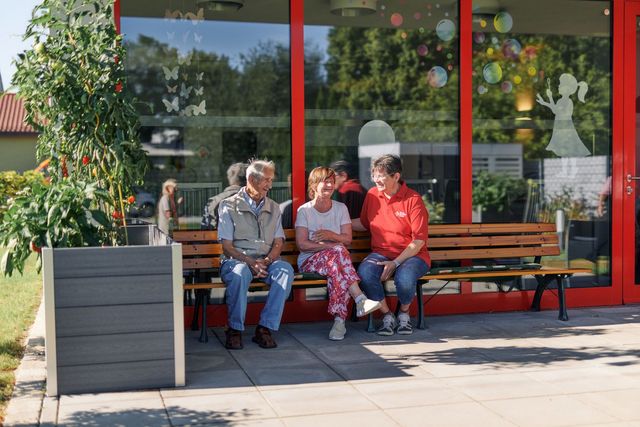 Mitarbeiterin des DRK sitzt mit Gästen der Tagespflege auf einer Bank im Garten. 