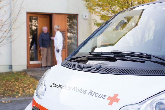 Mitarbeiterin einer Hauskrankenpflege steht mit Klienten vorm einem Haus.