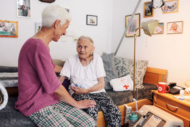 Mitarbeiterin des DRK sitzt mit Seniorin am Bett im Gespräch.