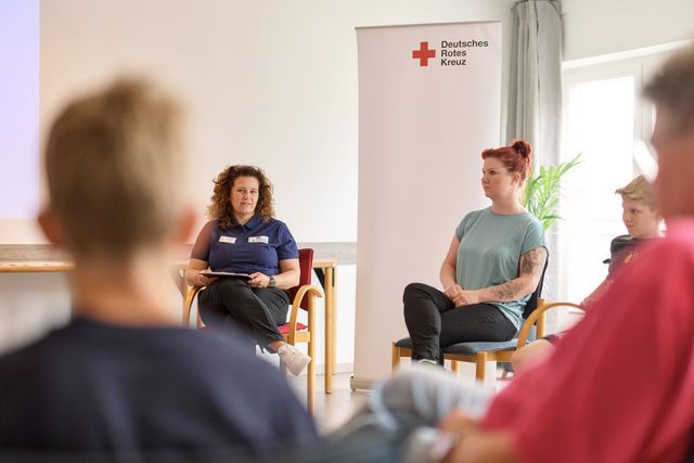 Eine Stuhlkreissituation mit Teilnehmenden und DRK-Mitarbeiter ist zu sehen.